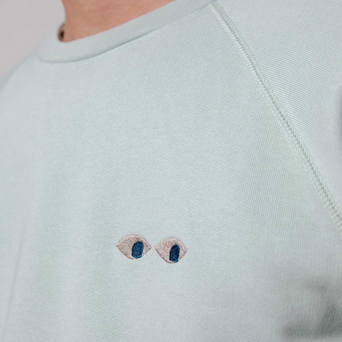 blassgrüner Pullover aus Bio-Baumwolle mit Stickerei auf der Brust: ein Comic Augenpaar.