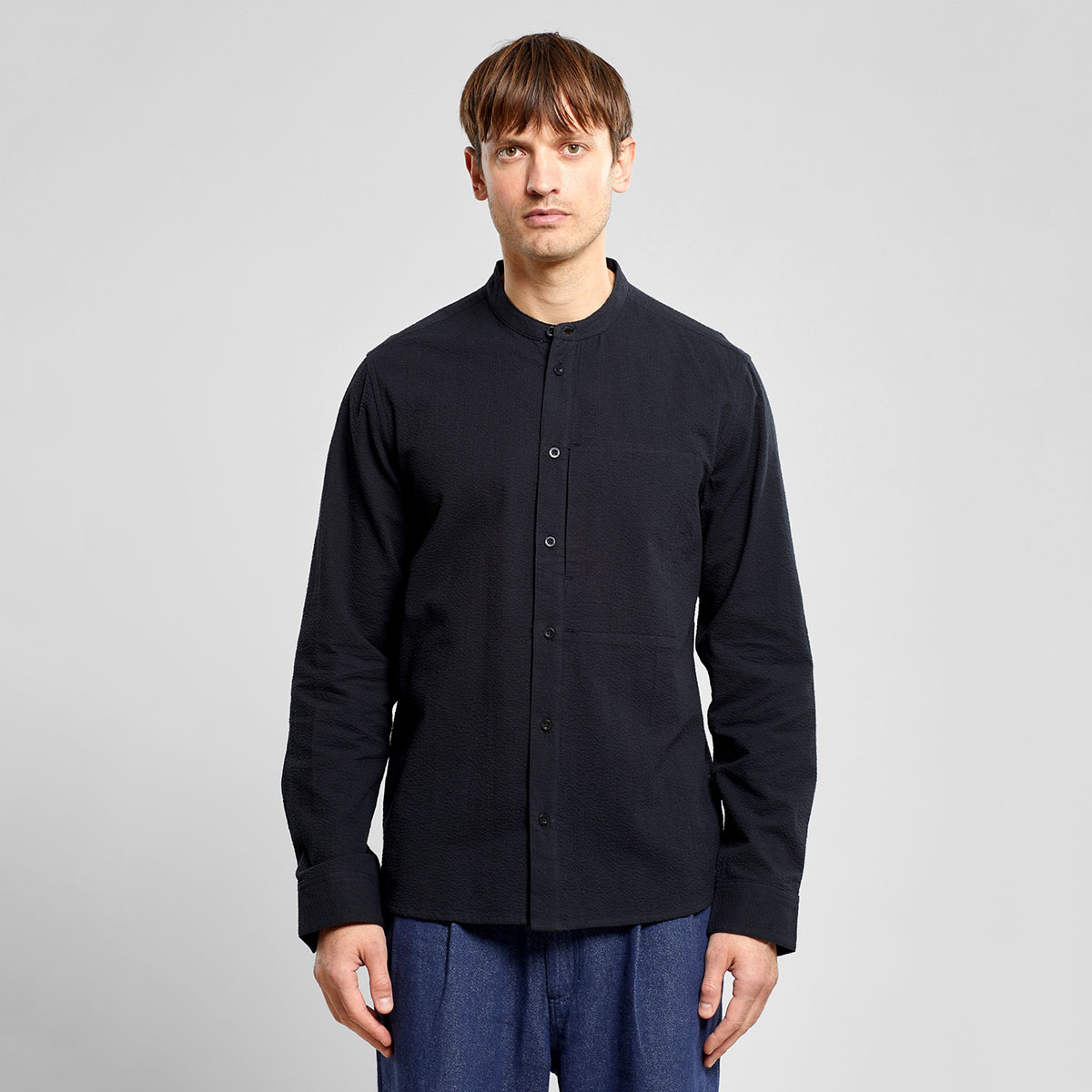 Schwarzes Hemd aus 100% Bio-Baumwolle in Seersucker Webung mit versetckter seitlicher Einschubtasche auf der linken Brustseite, Stehkragen und durchgehender Knopfleiste.
